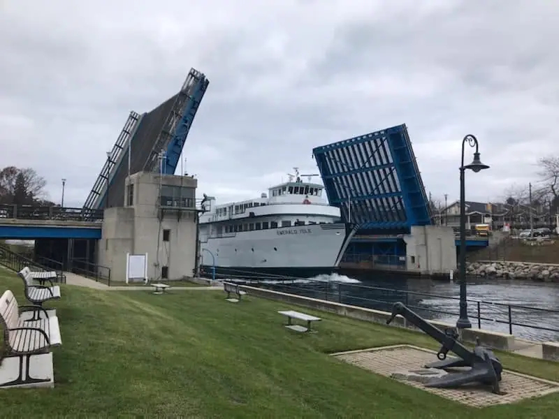 A white ferry passes through a raised drawbridge beside a grassy waterfront with benches and a large anchor.