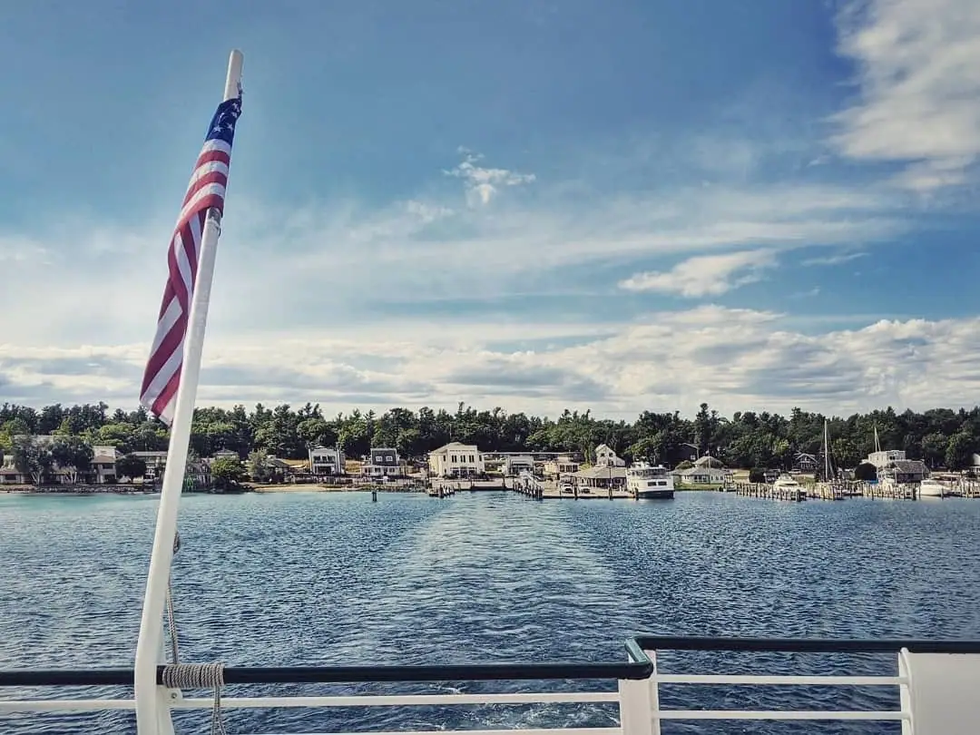 View from a boat stern toward a waterfront with docks, houses, trees, and an American flag.