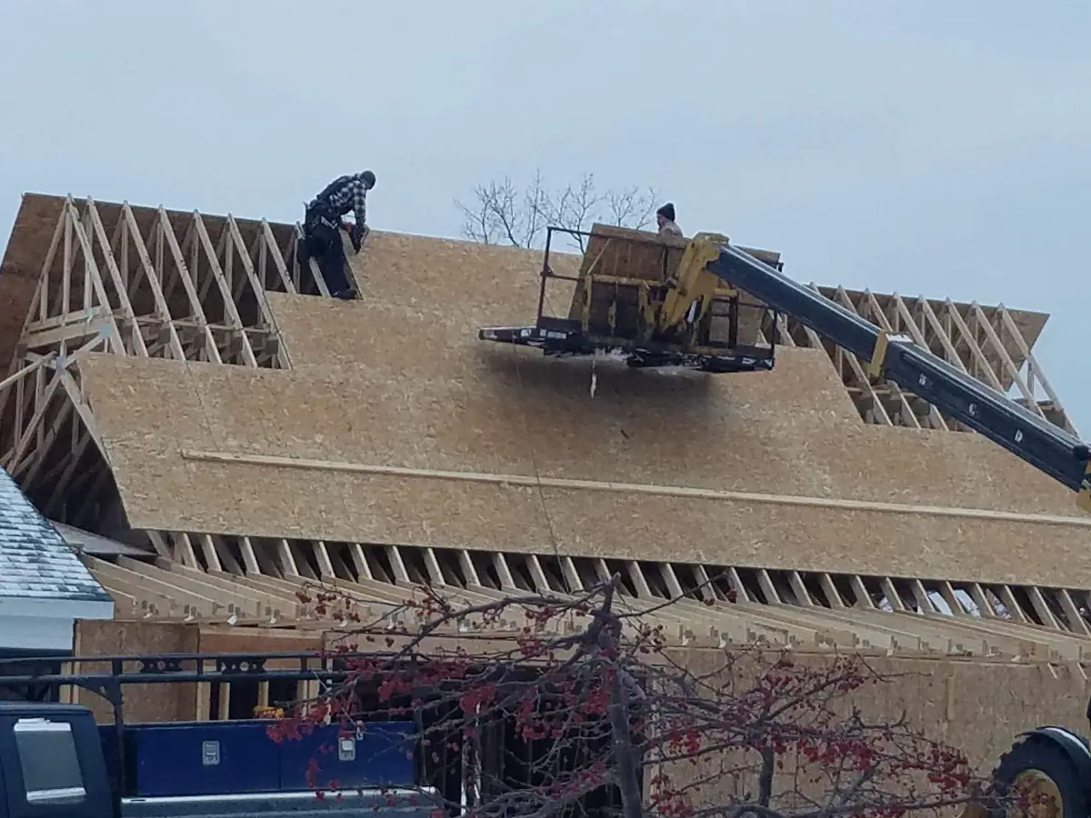Workers install sheathing on the roof of a timber-framed building using a boom lift.