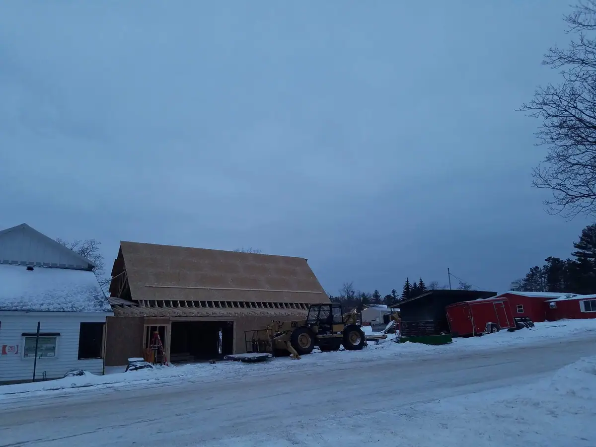 Snow-covered street beside a large building under construction, with a yellow loader and red trailers nearby.