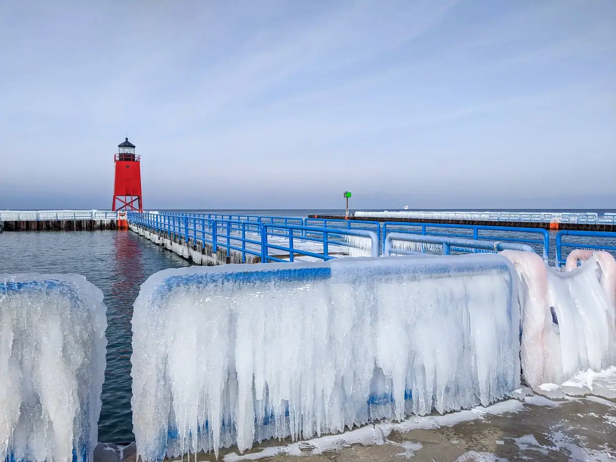 A red lighthouse and blue pier railings are coated in thick ice beside calm water.