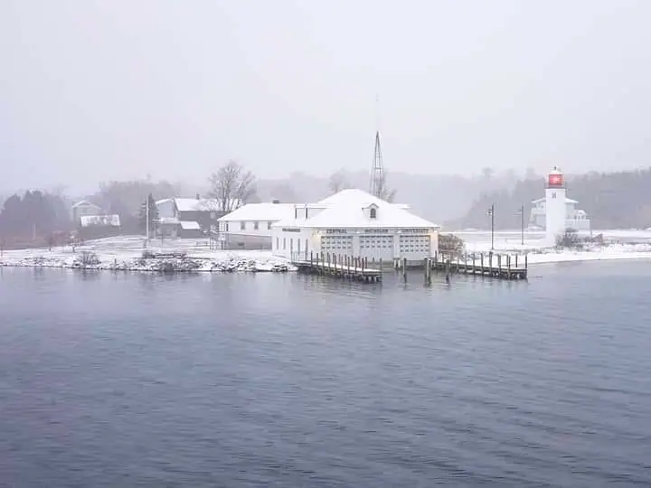 Snowy waterfront with a white building, pier, and lighthouse beneath a hazy winter sky.