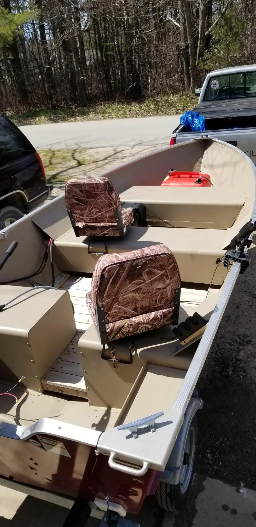 An aluminum boat with two camouflage seats rests on a trailer beside parked vehicles and a wooded roadside.