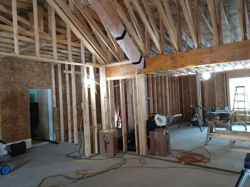 Interior construction scene with exposed framing, ductwork, tools, ladders, boxes, and cords on a concrete floor.