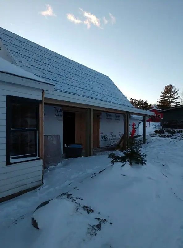 Snow-covered house under construction with Tyvek wrap, partial siding, and a small evergreen in the foreground.