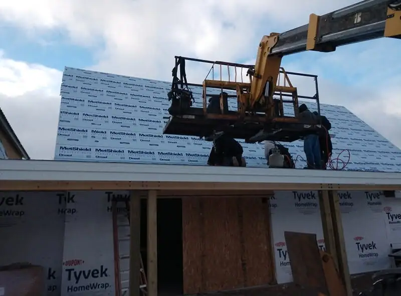 Workers use a boom lift while installing roof materials on a wood-framed building.