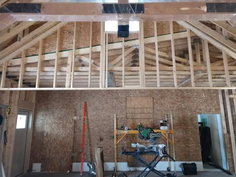 Interior of a partially framed building with exposed rafters, wall sheathing, plastic covering, and construction tools.