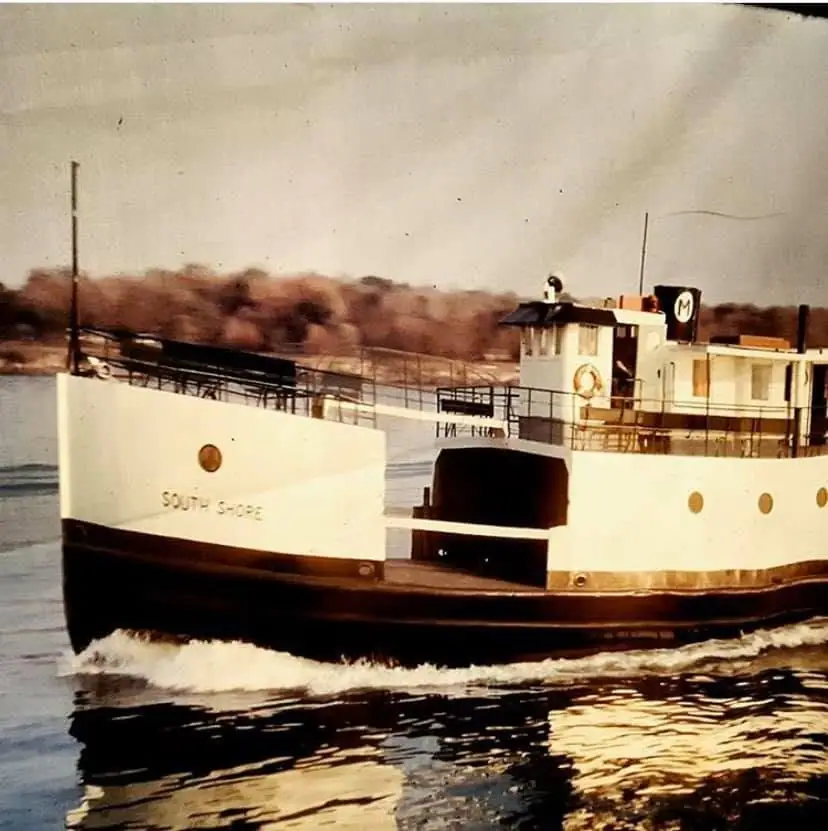 A white-and-black ferry named SOUTH SHORE moves through the water beside a wooded shoreline.