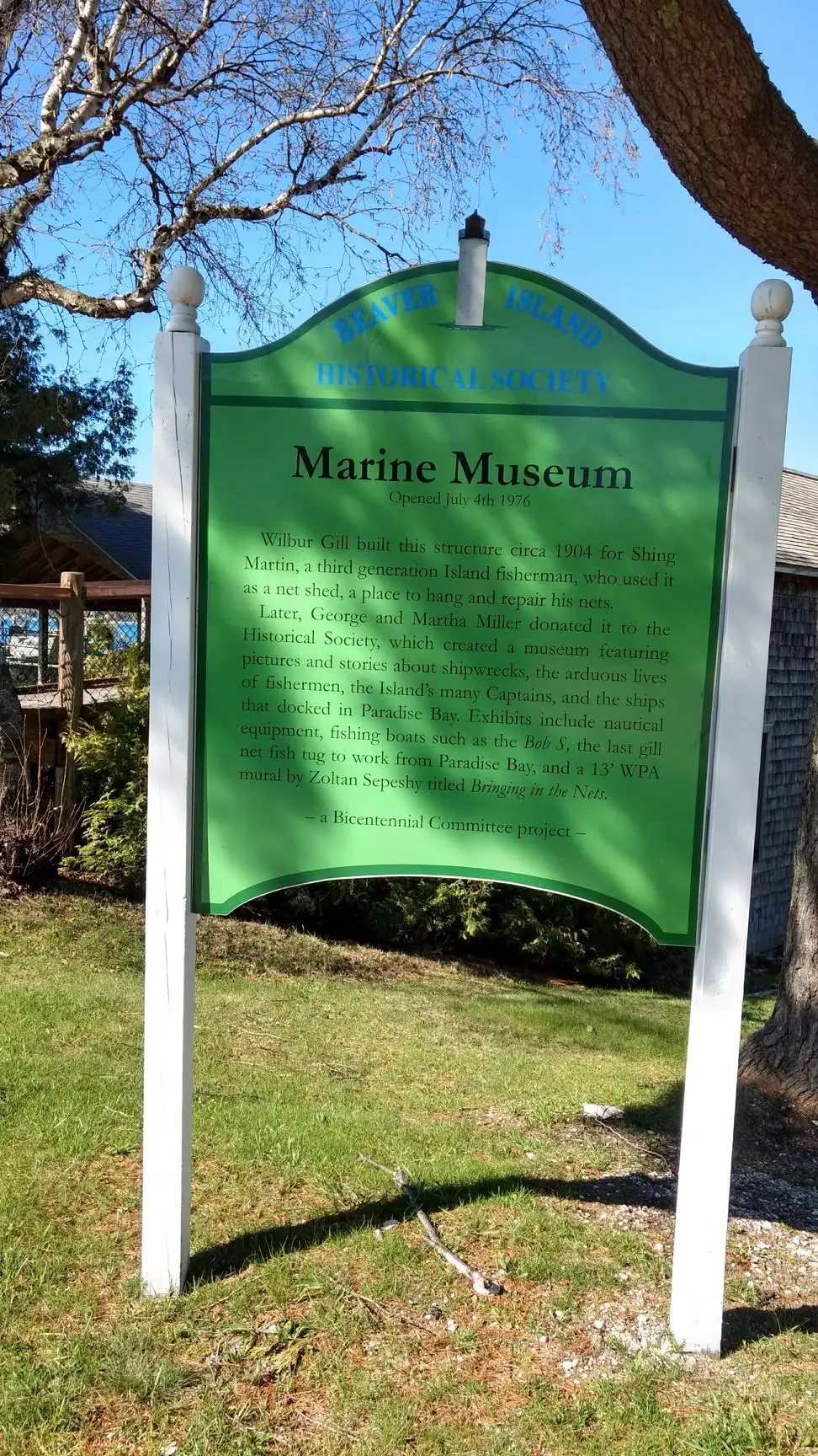 A green interpretive sign for a marine museum stands outdoors beneath leafless trees.