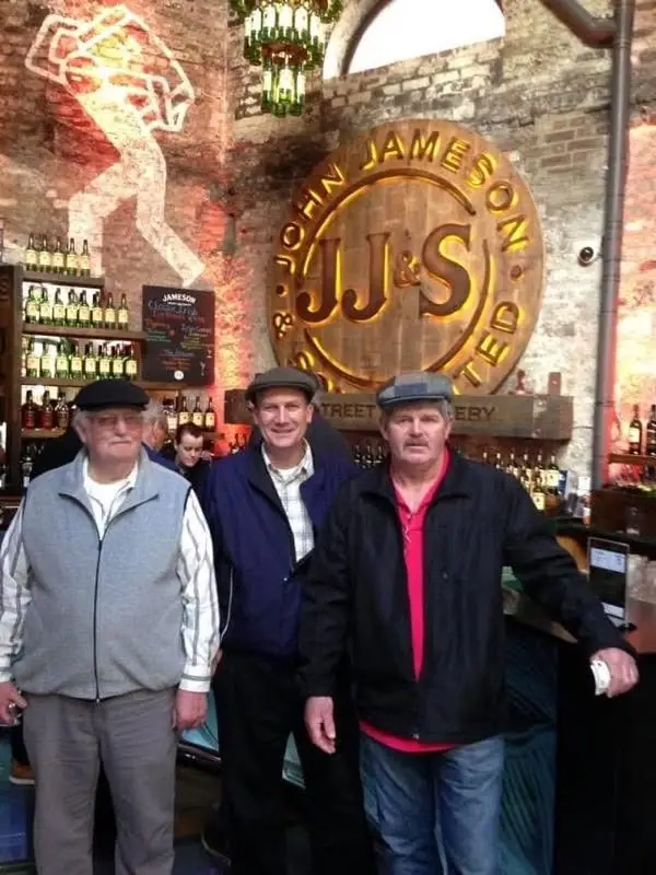 Three men stand inside a brick-walled bar beneath a large illuminated whiskey-company sign.