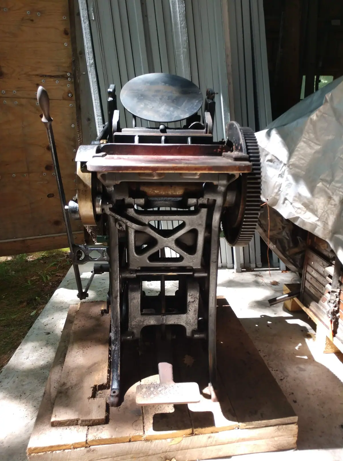 A cast-iron hand-cranked machine with large gears stands on a wooden platform in a shed or workshop.
