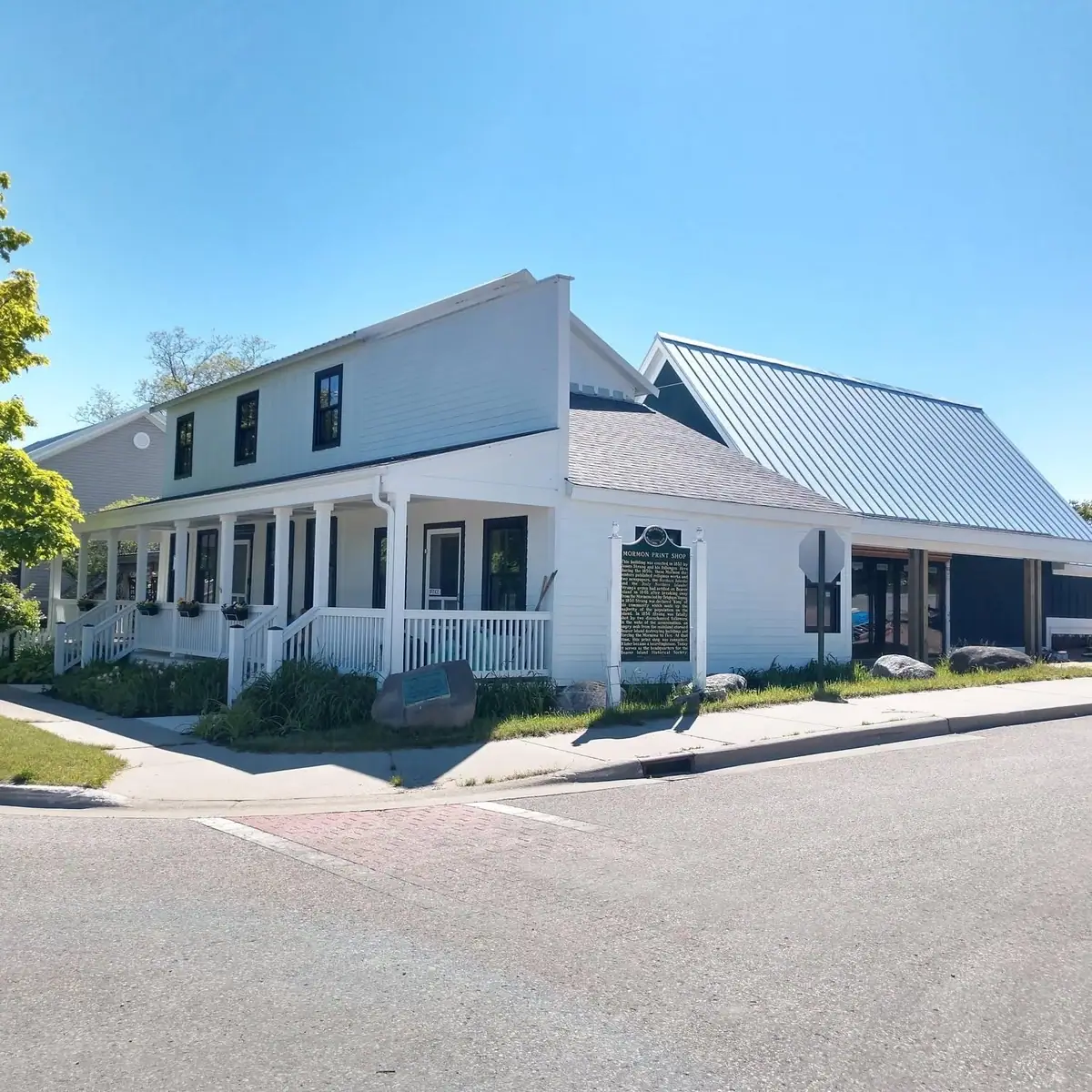 White two-story building with a porch, roadside marker, and adjacent metal-roofed structure.