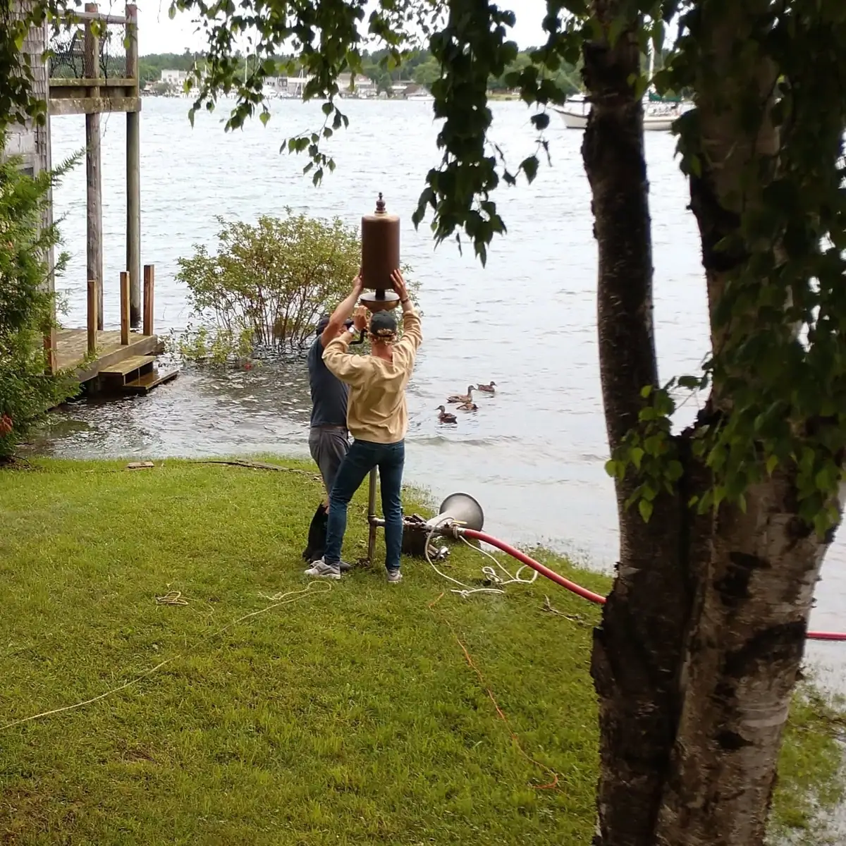 Two men hold a large cylindrical apparatus beside a waterfront, with ducks, a dock, and trees visible.