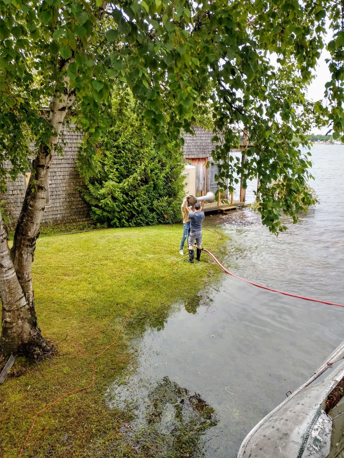 Two people handle a large horn-shaped apparatus beside a shingled waterside building and a red hose.