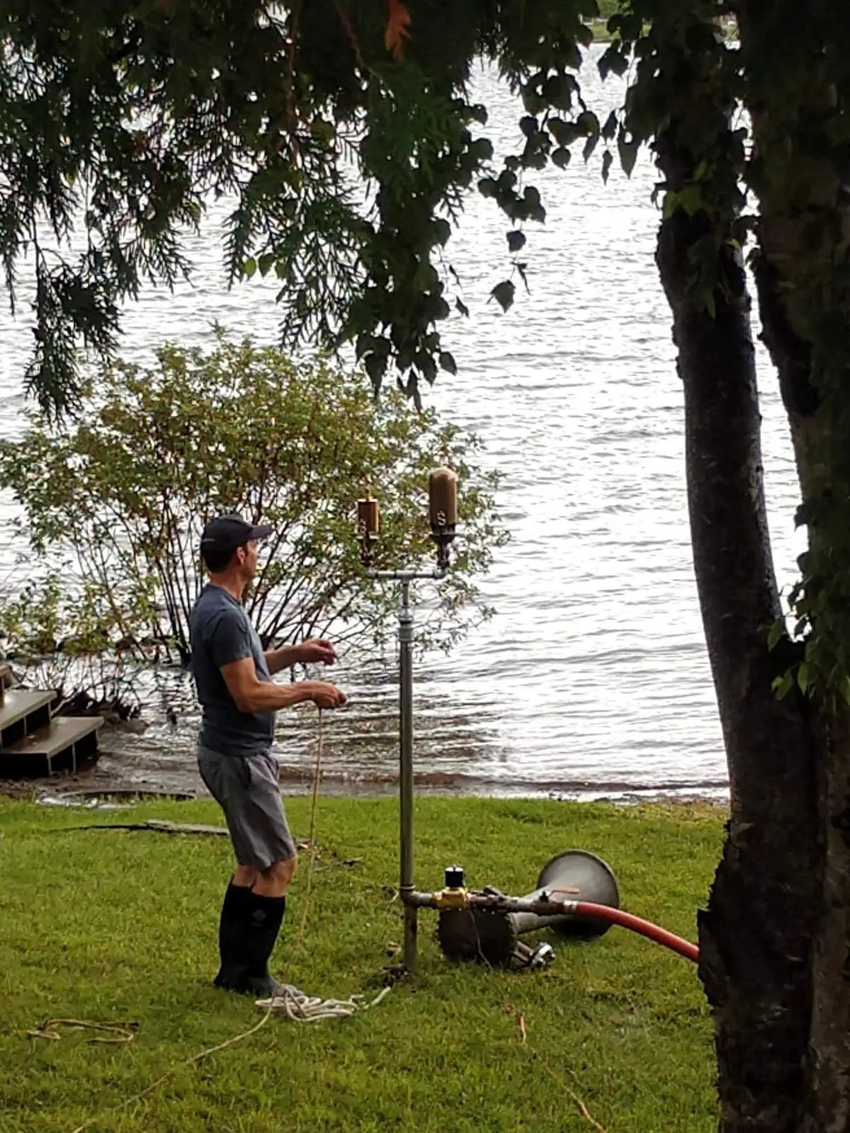 A man stands beside a metal apparatus with a red hose on a grassy shoreline.