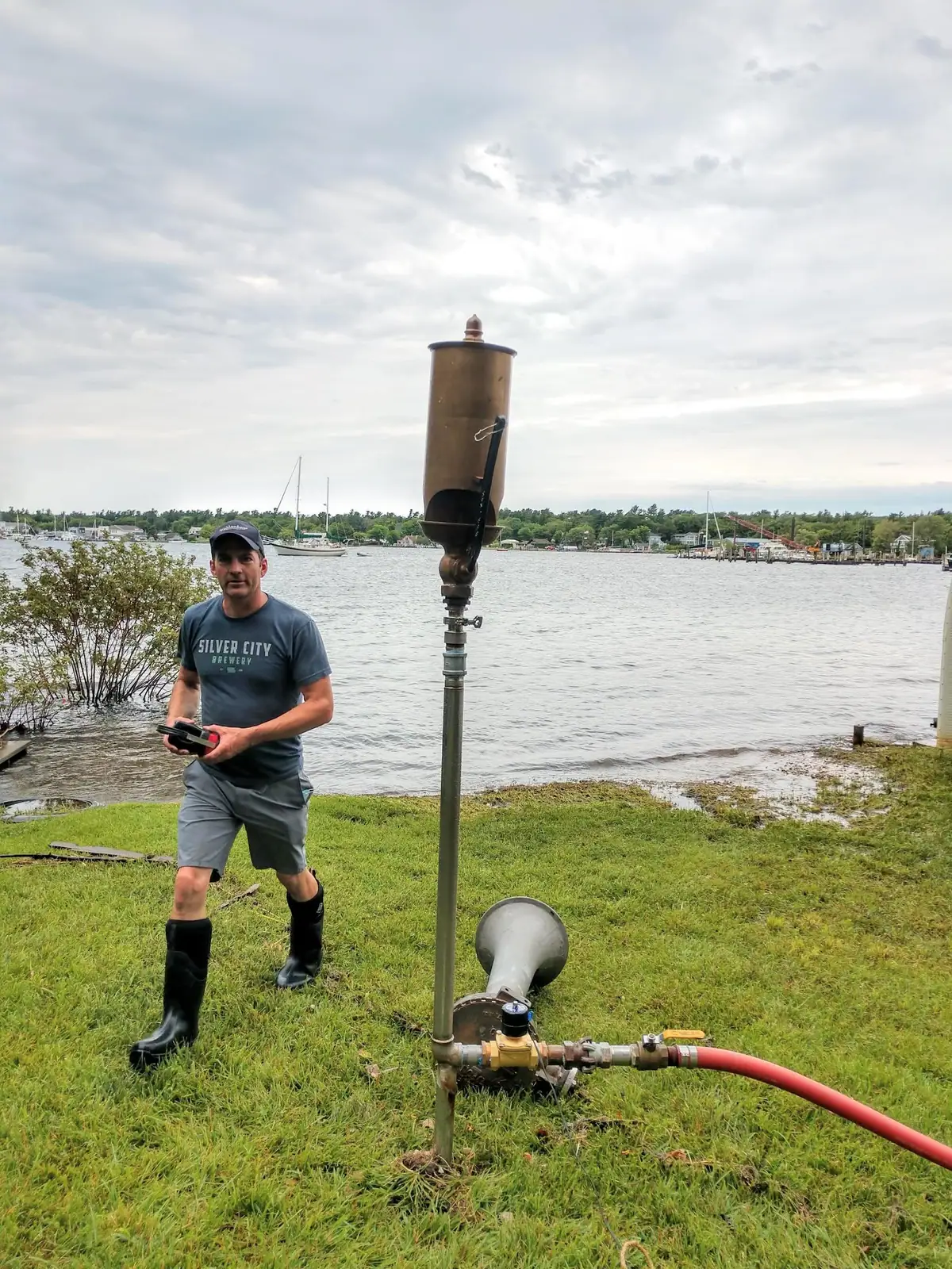 A man stands beside a tall metal whistle apparatus on a grassy waterfront with boats in the background.
