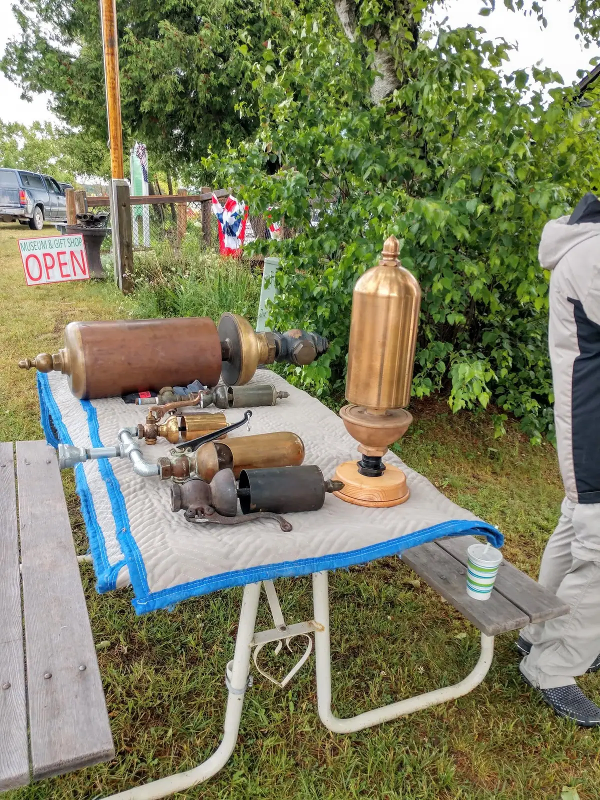 Outdoor table displaying antique whistle-like instruments beside a sign reading “MUSEUM & GIFT SHOP OPEN.”