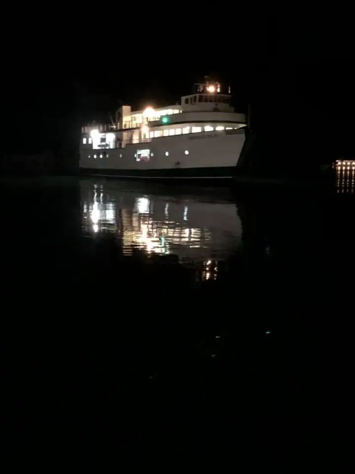 A brightly lit white passenger ferry moves across dark water at night, its lights reflected below.