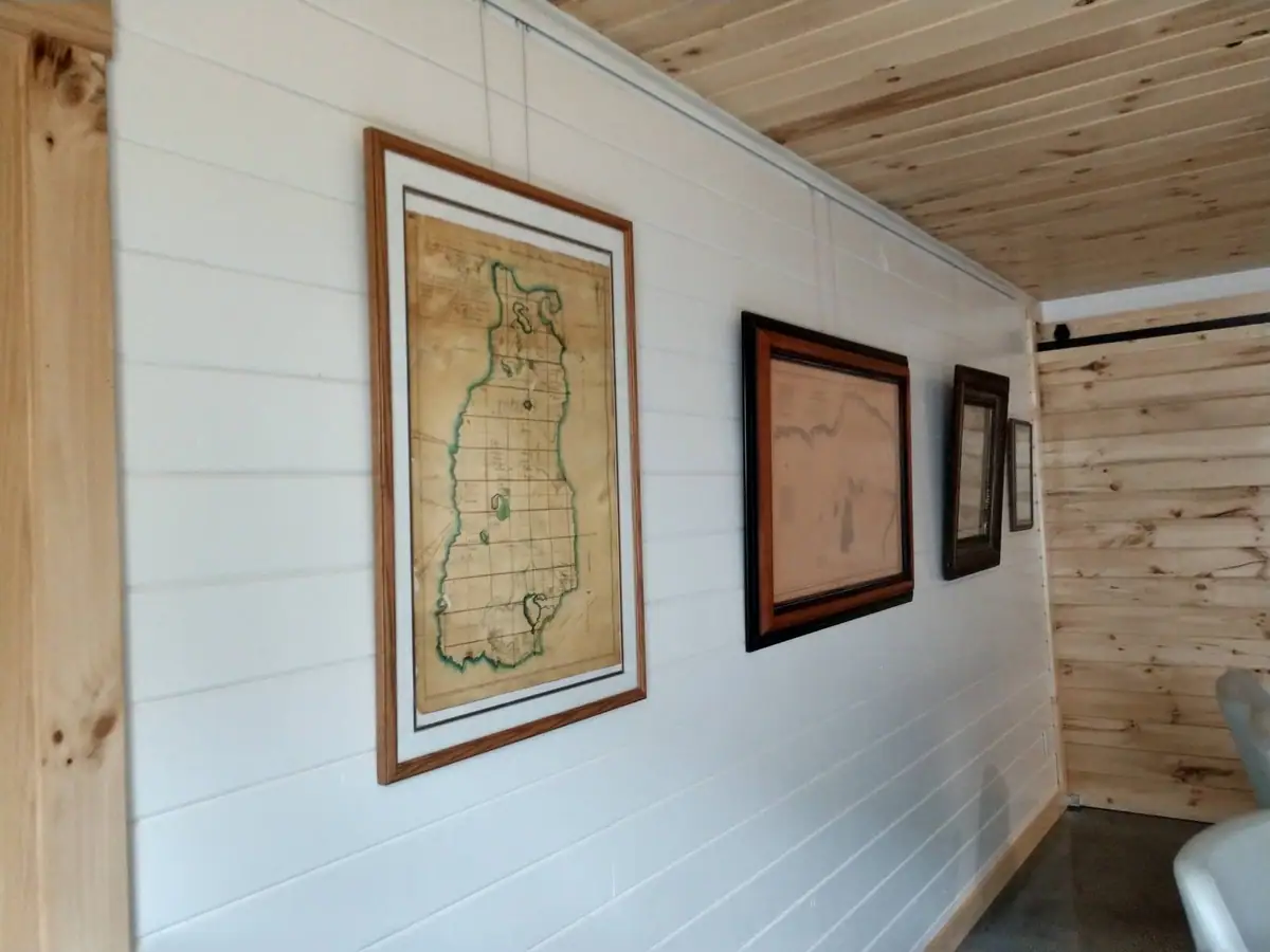Framed maps and documents hang on a white paneled wall in a wood-lined interior.