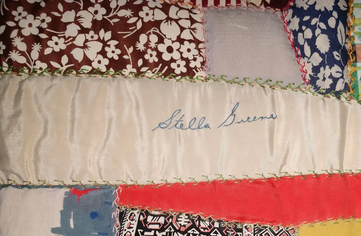 Close-up of a patchwork quilt with floral fabrics and blue embroidery reading “Stella Bucci.”