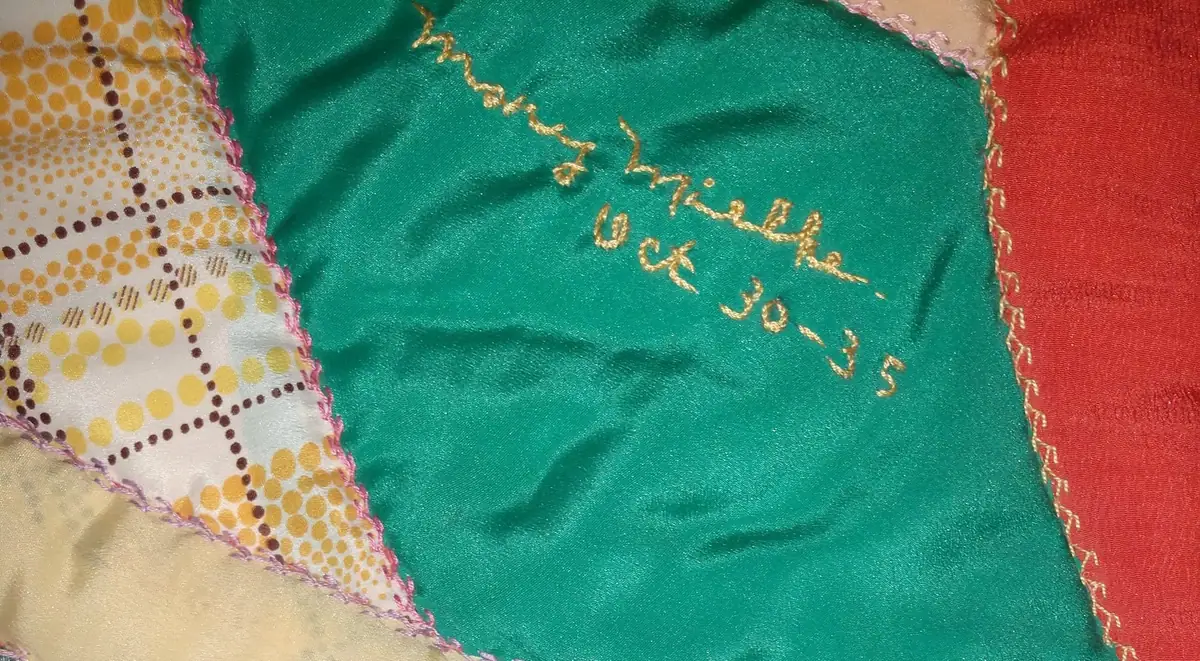 Close-up photograph of a colorful quilt with embroidered gold lettering and patterned fabric panels.