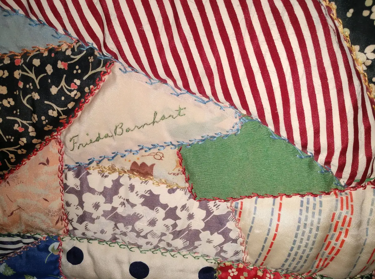 Close-up of a patchwork quilt with floral and striped fabric pieces and the embroidered name “Frieda Barnhart.”