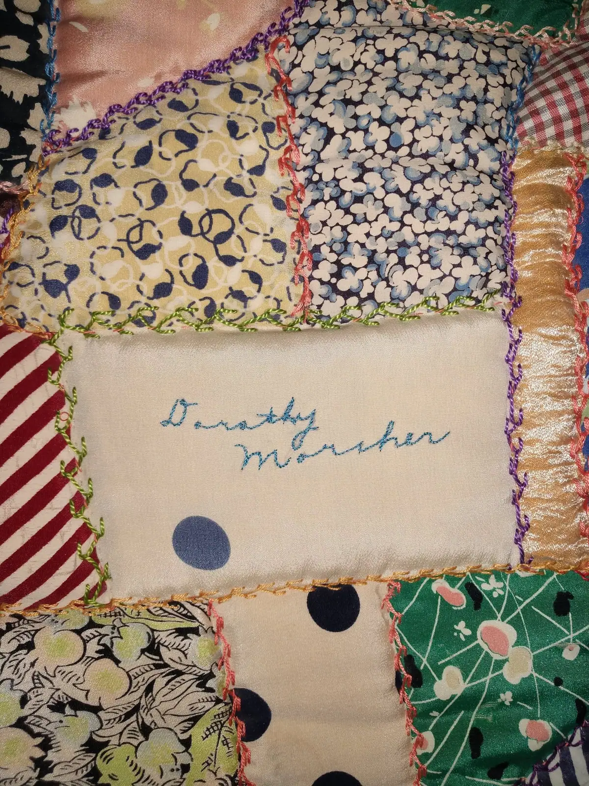Close-up photograph of a colorful patchwork quilt with embroidered lettering and contrasting stitched fabric borders.