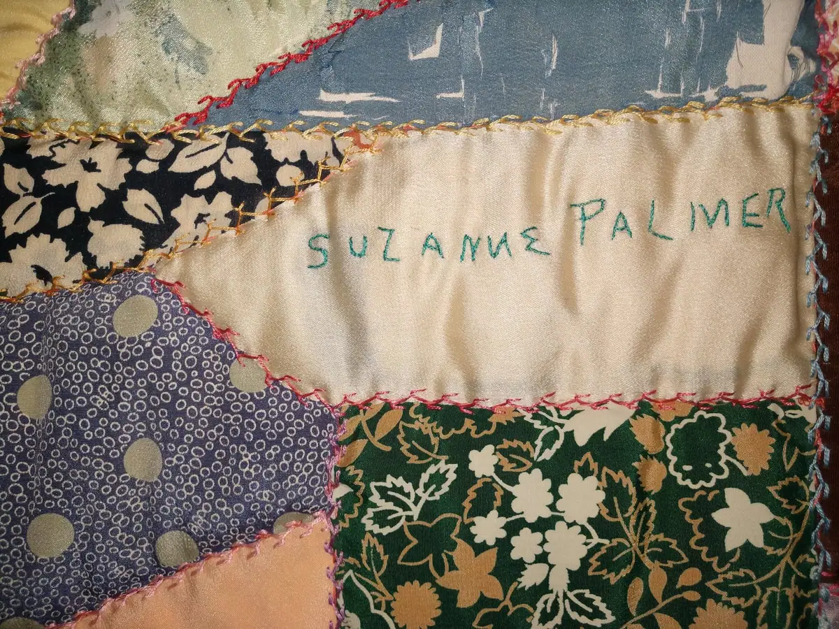 Close-up photograph of a colorful hand-stitched patchwork quilt bearing the embroidered name “SUZANNE PALMER.”