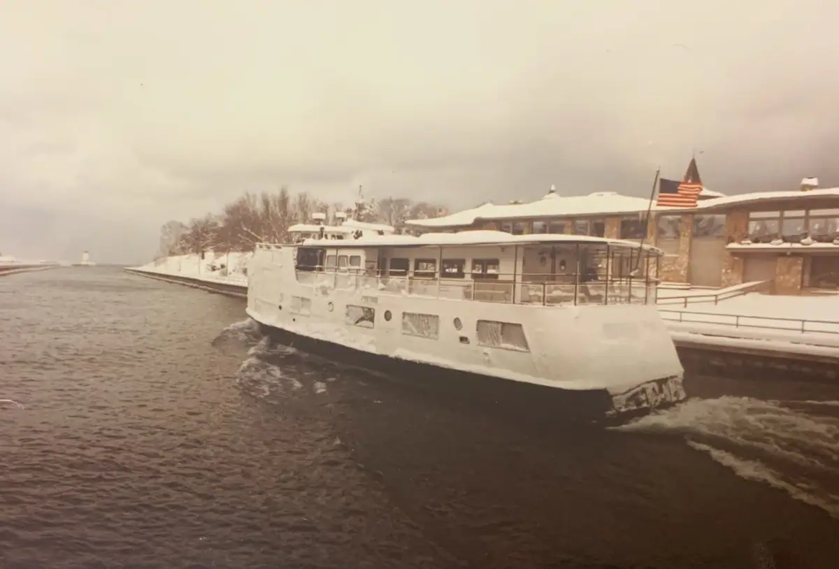 A snow-covered white passenger ferry moves beside a waterfront terminal under a cloudy winter sky.