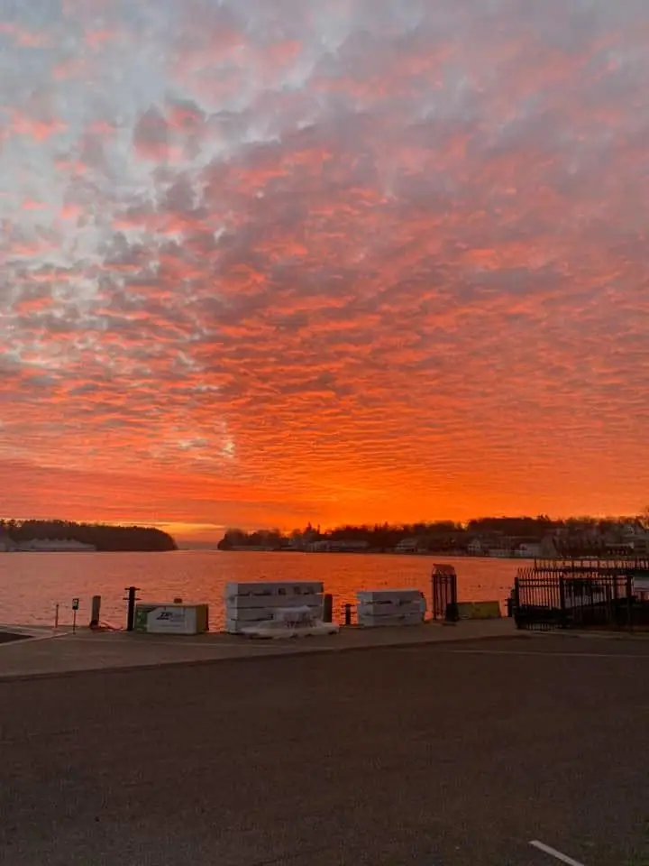 Orange-red sunset over calm water, with dark shorelines and a paved waterfront in the foreground.