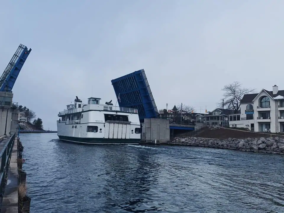 A white ferry passes beneath a raised blue drawbridge along a developed waterfront.