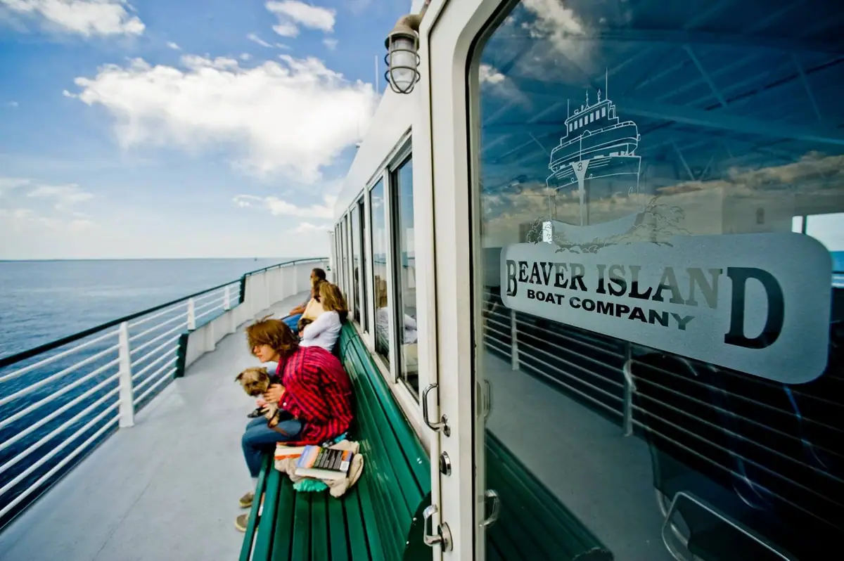 Passengers sit with a dog on a ferry deck beside a glass door marked with a company logo.