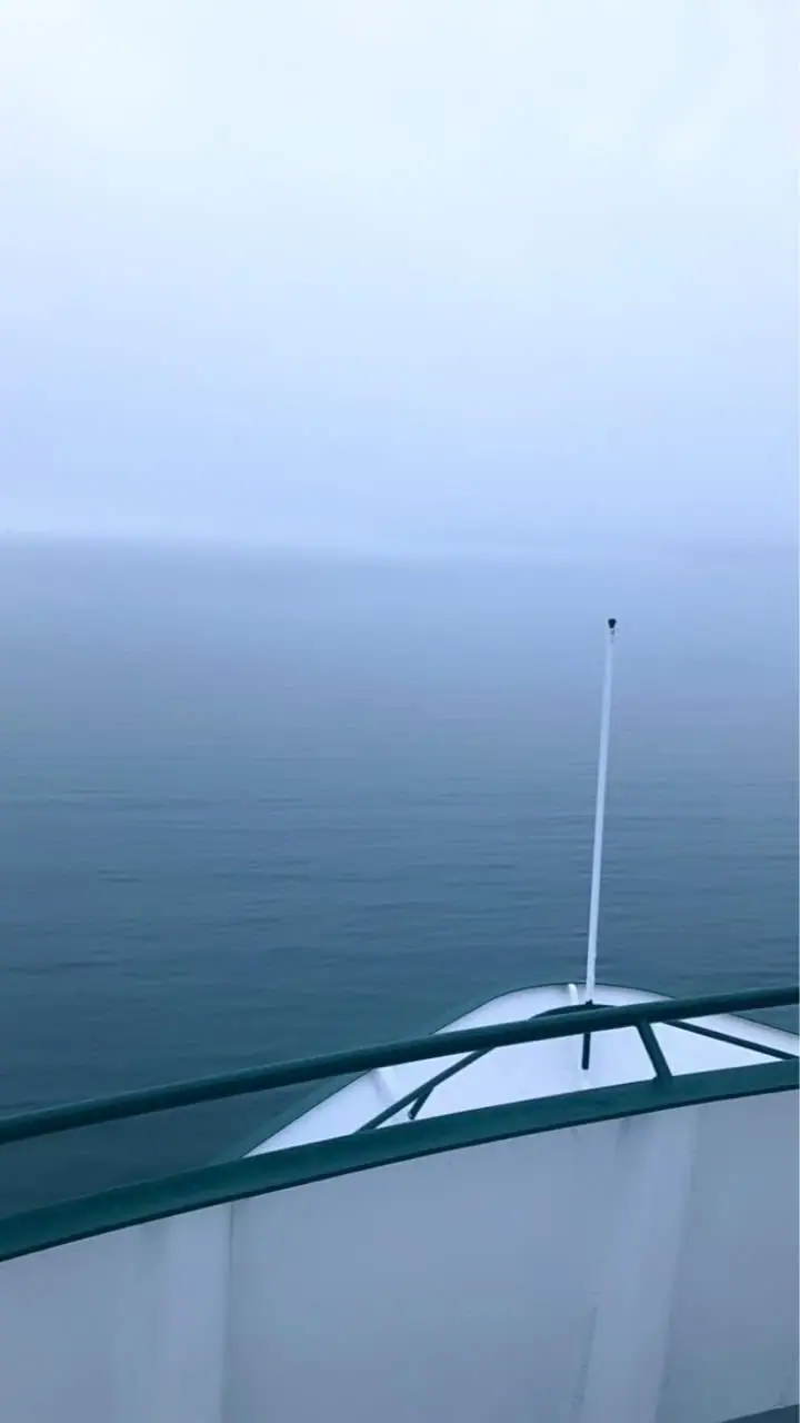 View over calm water from a vessel’s forward deck, with railings and a slim antenna in the foreground.