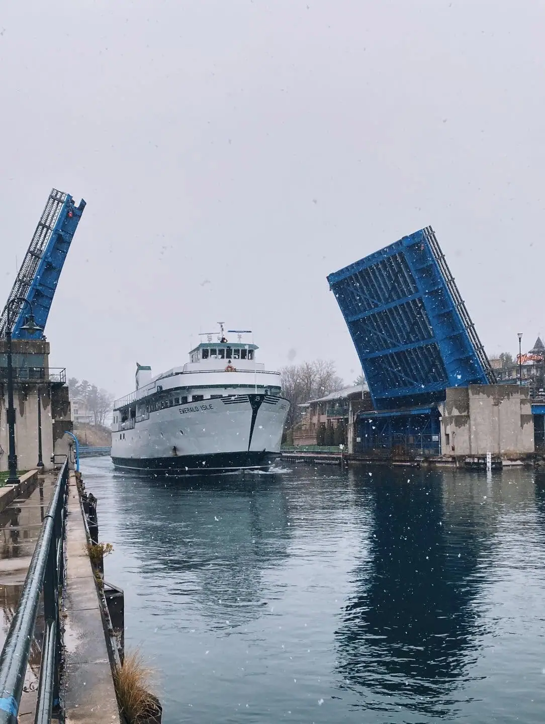 A white ferry passes between two raised blue drawbridges as snow falls.