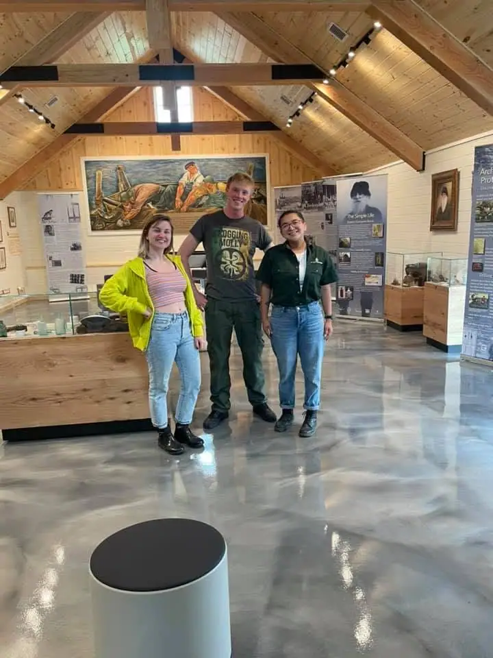 Three people pose inside a wood-paneled exhibition hall with display cases and a large painting behind them.