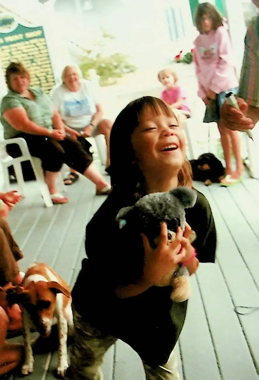 Laughing child holding a plush koala on a porch while adults, children, and dogs gather nearby.