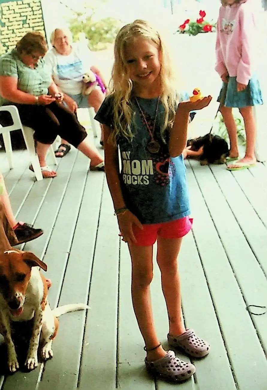 A smiling girl holds a yellow rubber duck on a wooden deck while people and dogs appear behind her.
