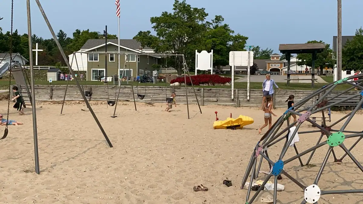 Children and adults use a sandy playground with swings, climbing equipment, and a flagpole in the background.