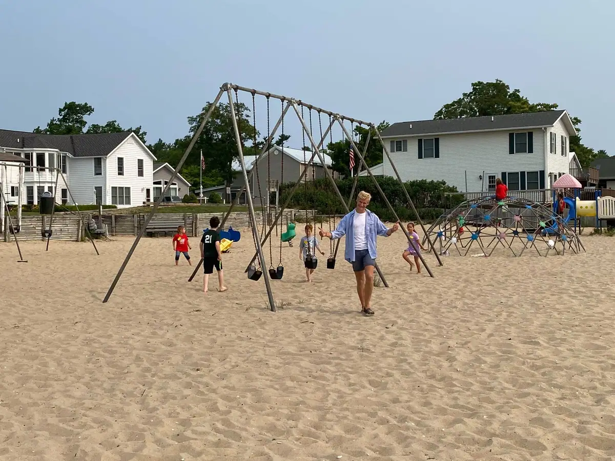 Adults and children gather on a sandy playground with swings and climbing equipment in front of houses.