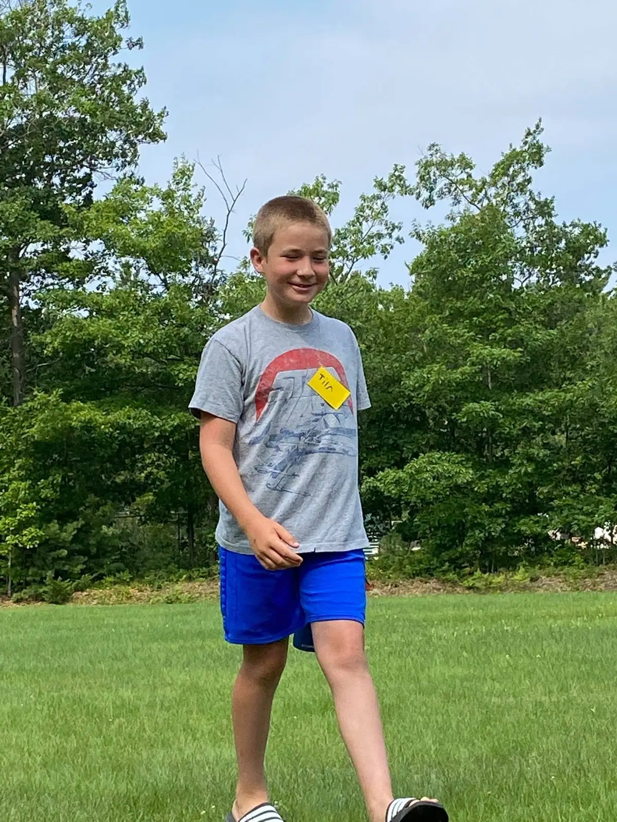 A smiling boy wearing a yellow name tag stands in a grassy field with trees behind him.