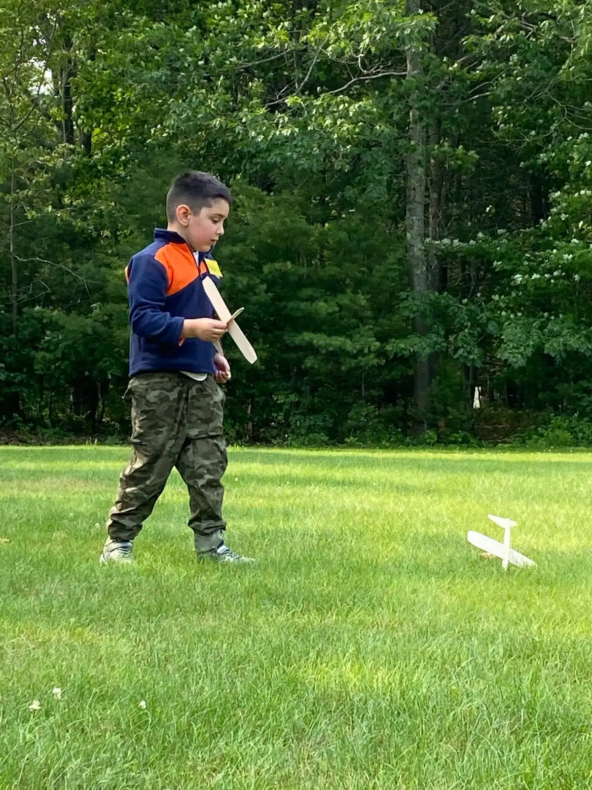 A child stands on a grassy lawn holding a wooden model airplane, with another model airplane on the ground nearby.