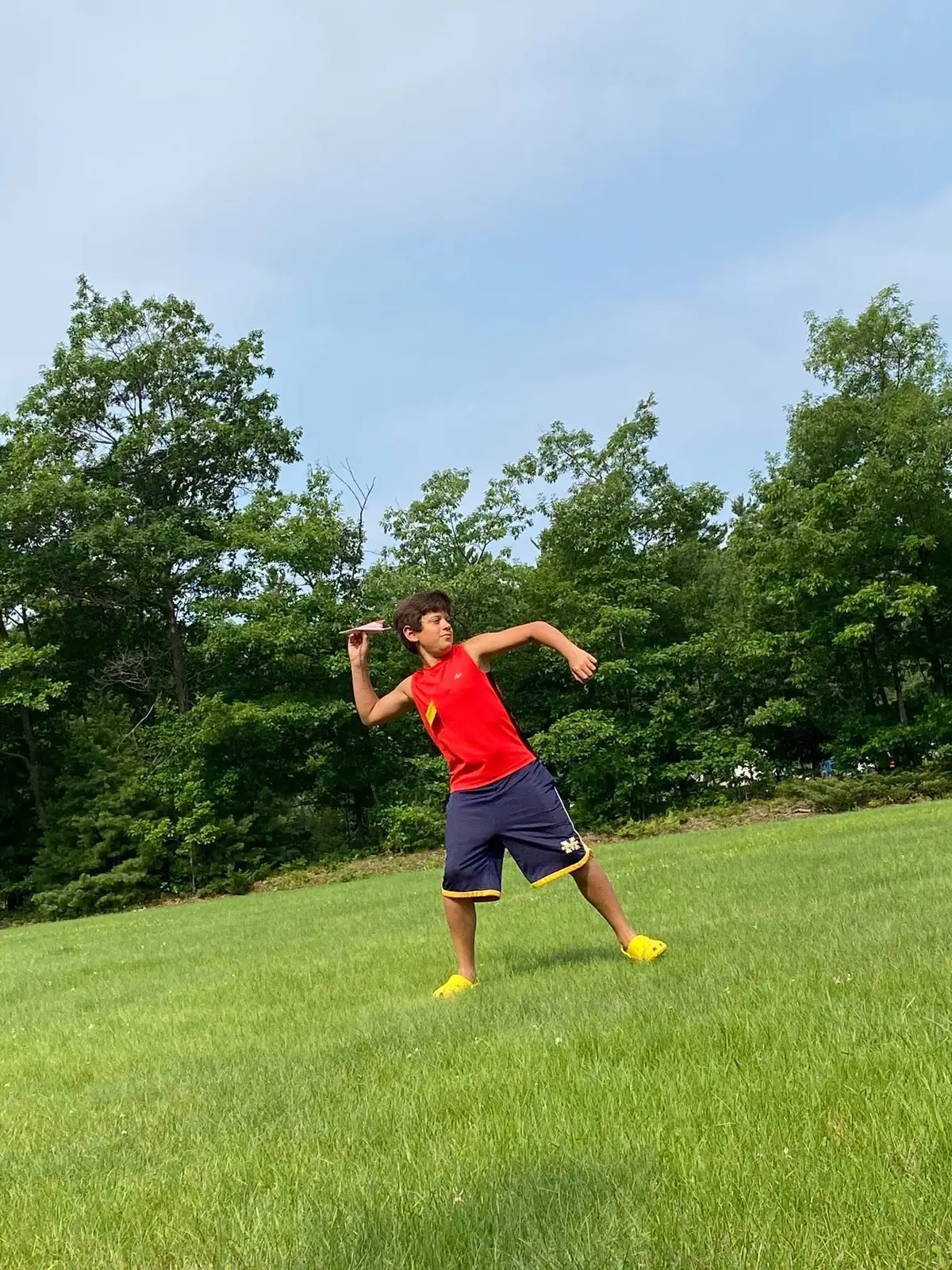 Child in a red shirt throws a pink paper airplane across a grassy field.