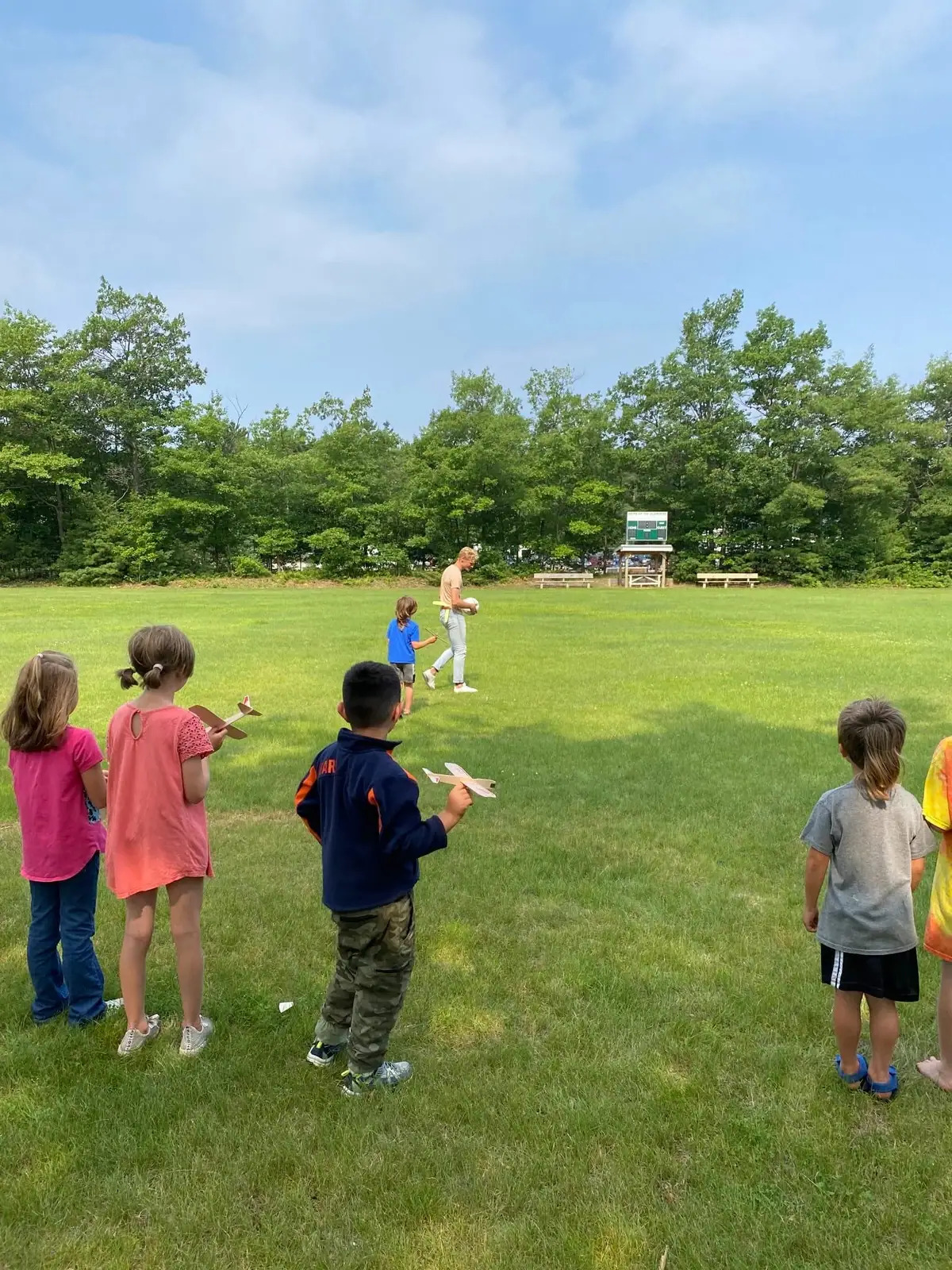 Children hold paper airplanes while an adult stands in a grassy field beside a scoreboard and benches.