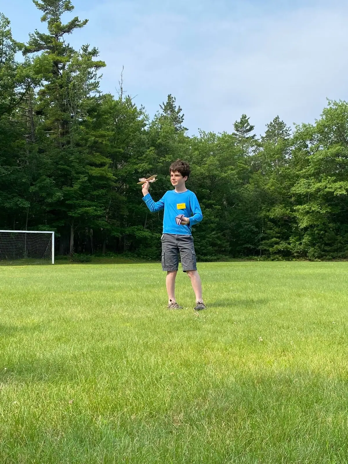 A child holds a small toy airplane in a grassy field beside a soccer goal and trees.