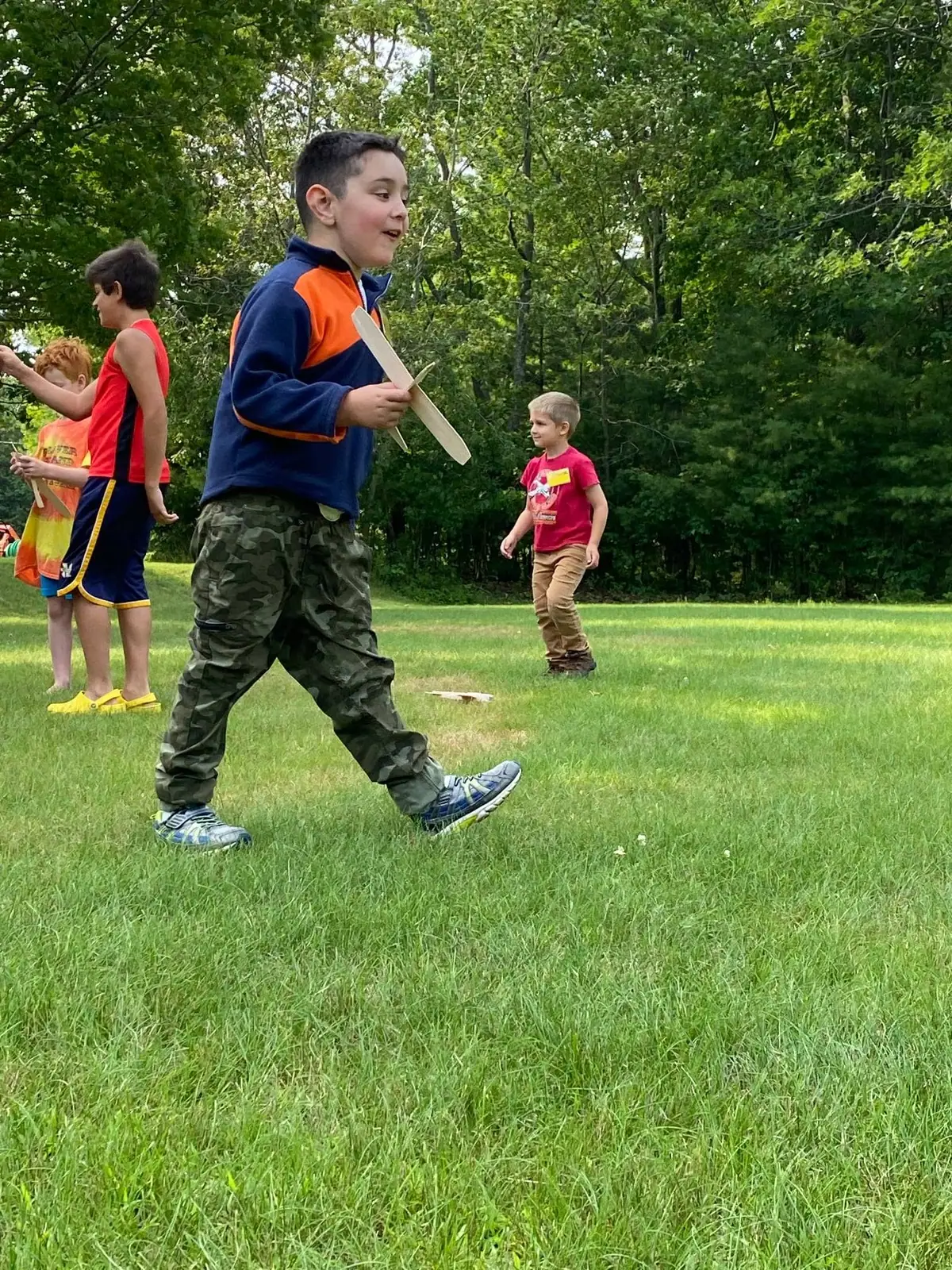 Four boys play with wooden model airplanes on a grassy lawn beside a wooded area.