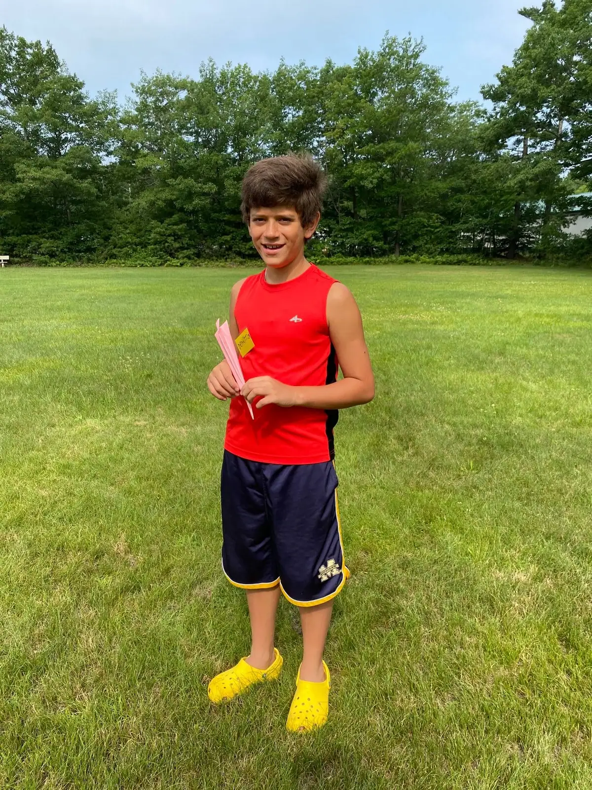 A boy in athletic clothing stands in a grassy field holding pink papers and a small yellow tag.