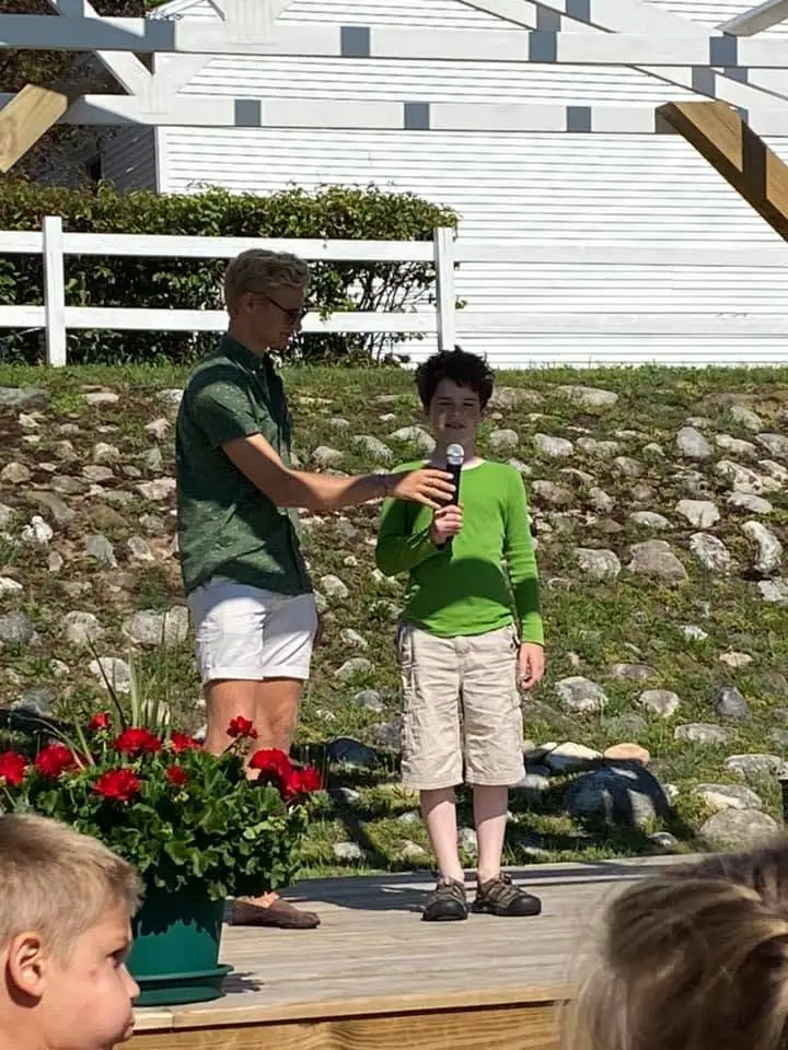An adult holds a microphone toward a child on a wooden platform beside a flower pot and white fence.