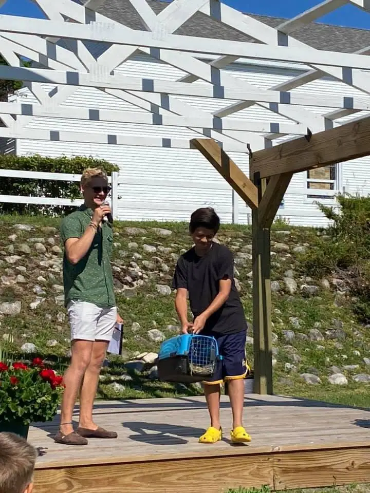 Two people stand on a wooden deck beneath a pergola, with one holding a microphone and the other holding a pet carrier.