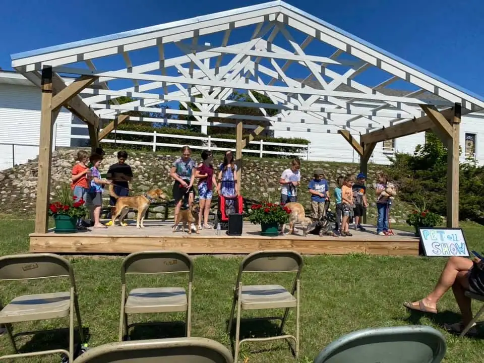 Children and dogs gather on a decorated outdoor stage beneath a white lattice canopy during a pet show.