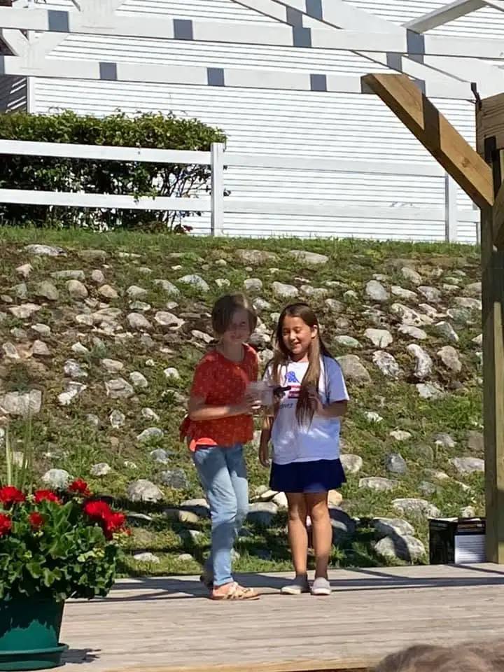 Two girls stand on a wooden deck beside potted flowers, with a rocky slope and white-sided building behind them.